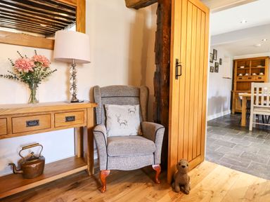 A corner with a gray armchair and a pillow with deer designs next to a wooden table with a lamp and flowers and a stuffed animal on wooden floor at Cwm Yr Afon Mawr in Llanbedr near Harlech