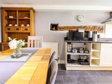 A kitchen with a wooden dining table and chairs a flower vase a wooden cabinet kitchen counter with microwave pots pans and knives on the counter at Cwm Yr Afon Mawr in Llanbedr near Harlech