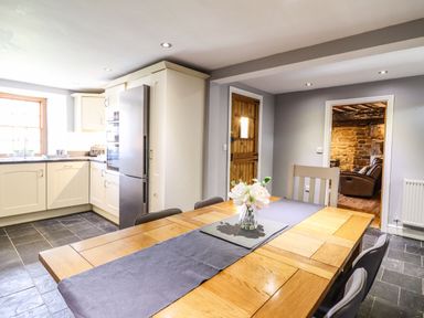 A kitchen with a wooden dining table and chairs a vase with flowers on the table a stainless steel fridge and cream cabinets at Cwm Yr Afon Mawr in Llanbedr near Harlech