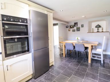 A kitchen with a double oven, a refrigerator, a wooden dining table with chairs, and framed pictures on the wall at Cwm Yr Afon Mawr in Llanbedr near Harlech