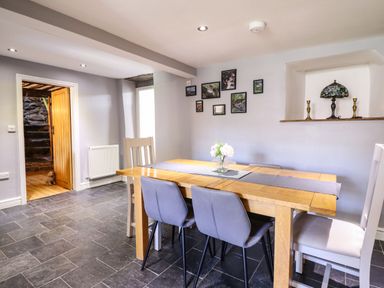 A dining room with a wooden table and mixed chairs with a vase of flowers on the table wall with framed pictures and a recessed shelf with a lamp and candlesticks at Cwm Yr Afon Mawr Llanbedr near Harlech