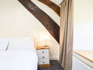 A bedroom with a white bed next to a bedside table with a lamp under a wooden beam and beige curtains at Cwm Yr Afon Mawr in Llanbedr near Harlech