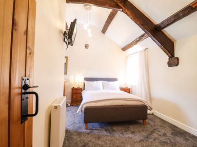 A bedroom with a double bed, wooden bedside tables, a wall-mounted TV, exposed wooden beams on the ceiling, and light coming through a window at Cwm Yr Afon Mawr in Llanbedr near Harlech