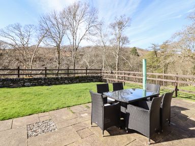 An outdoor patio area with a glass table and six chairs surrounded by trees and a wooden fence at Cwm Yr Afon Mawr in Llanbedr near Harlech