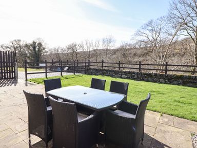 An outdoor patio with a glass table and six wicker chairs next to a grass area at Cwm Yr Afon Mawr in Llanbedr near Harlech