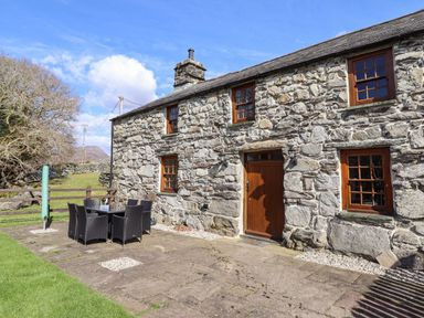 A stone cottage with wooden windows and a door on a patio with outdoor dining furniture at Cwm Yr Afon Mawr in Llanbedr near Harlech