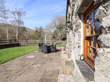 A stone building with a wooden window next to a paved patio with outdoor dining furniture and a green lawn at Cwm Yr Afon Mawr in Llanbedr near Harlech
