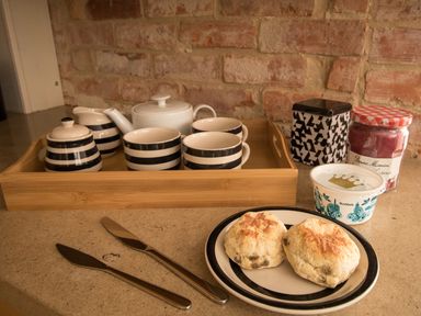 A kitchen counter with a wooden tray holding a teapot cream and sugar containers cups a plate with two scones two butter knives a jar of jam and a container of clotted cream at Sandsfoot Rest in Weymouth