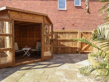 A wooden garden shed with open doors showing two chairs and a small table inside in a paved backyard at Sandsfoot Rest in Weymouth