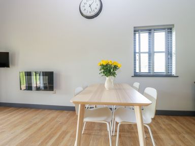 A dining area with a wooden table four white chairs a vase of yellow flowers on the table a wall clock and a window with blinds at Foxglove in Sticklinch West Pennard Glastonbury