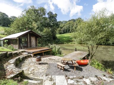 A small wooden cabin near a pond with a wooden deck and a stone patio with a fire pit and picnic table by Foxglove in Sticklinch West Pennard Glastonbury
