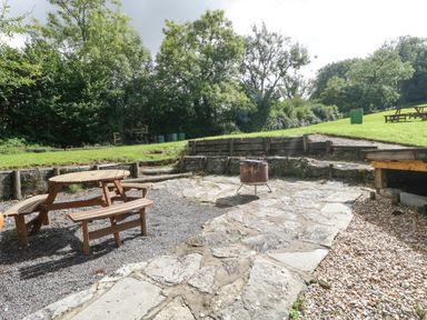 A patio area with a wooden picnic table and fire pit surrounded by grass and trees at Foxglove in Sticklinch West Pennard Glastonbury