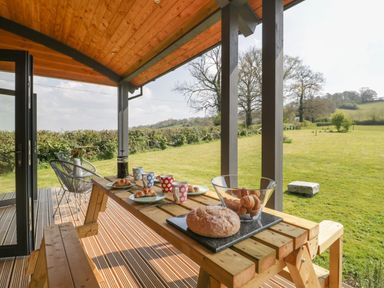 A wooden picnic table with bread and croissants on a covered patio overlooking a grassy field at Foxglove in Sticklinch West Pennard Glastonbury