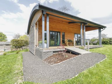 A wooden house with a sloped roof and a ramp leading to a porch with chairs and a bench surrounded by grass at Foxglove in Sticklinch West Pennard Glastonbury