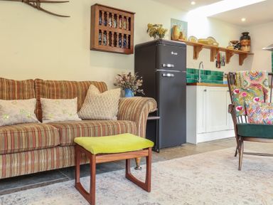 A living area with a striped sofa beside a green cushioned stool and a dark fridge near a kitchen counter at Courtyard Cottage Poplar Farm Barn Sweffling near Saxmundham