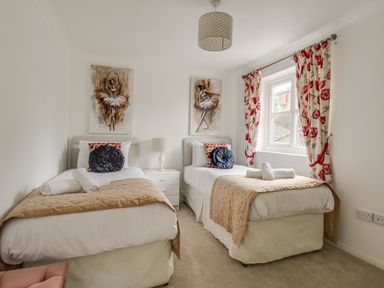 A bedroom with two single beds with beige covers and decorative pillows a window with floral curtains and a bedside table with a lamp at Casa Carmen in Weymouth