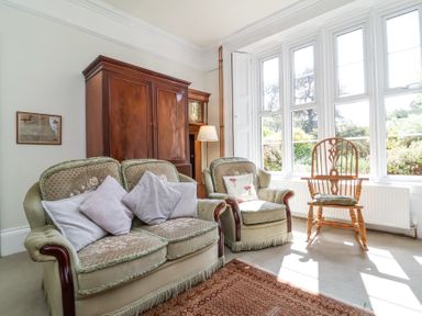 A sitting room with two upholstered armchairs a wooden cabinet a standing lamp a wooden chair and large windows at The Old Vicarage in Tiverton