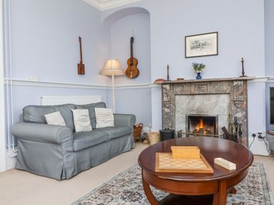 A living room with a gray sofa white pillows a wooden coffee table with a game board a fireplace with a fire two guitars on the wall and a floor lamp at The Old Vicarage in Tiverton