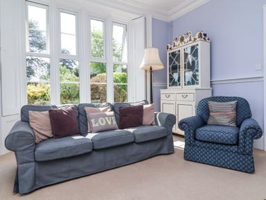 A sitting room with a sofa and armchair near large windows and a cabinet at The Old Vicarage in Tiverton