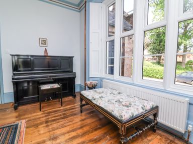 A room with a black upright piano and floral cushioned bench near large windows at The Old Vicarage in Tiverton