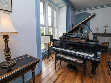 A room with a black grand piano a piano bench a music stand with sheet music a table lamp and a fireplace at The Old Vicarage in Tiverton