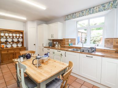 A kitchen with a wooden table and chairs a cabinet with plates a window and white cabinets at The Old Vicarage in Tiverton
