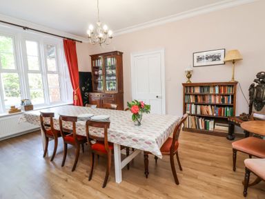 A dining room with a long table covered with a tablecloth surrounded by wooden chairs near a window and bookshelves at The Old Vicarage in Tiverton