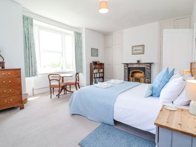 A bedroom with a bed blue bedding wooden side table dresser small dining table and chairs bookshelf and fireplace at The Old Vicarage in Tiverton