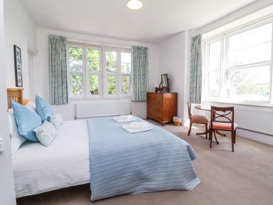 A bedroom with a double bed covered with a blue quilt and pillows two windows with patterned curtains a wooden chest of drawers and a small round table with two chairs at The Old Vicarage in Tiverton