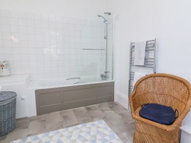 A bathroom with a bathtub and shower, a wicker chair with cushion, a towel rack with towels, and a laundry basket at The Old Vicarage in Tiverton