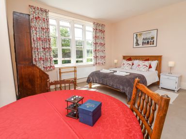 A bedroom with a double bed pillows and bedside tables near a window with floral curtains and a table with a red cloth and wooden chairs at The Old Vicarage in Tiverton