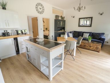 An open-plan kitchen and living area with a kitchen island, dining table with chairs, sofa, coffee table, and wall clock at The Barn in Droitwich Spa
