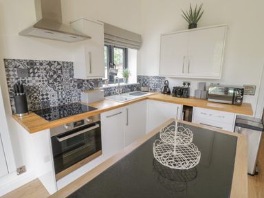 A kitchen with wooden countertops, white cabinets, patterned gray backsplash, oven, sink, toaster, kettle, microwave, and a two-tiered wire fruit basket at The Barn in Droitwich Spa