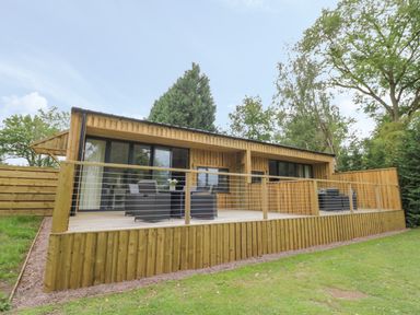 A wooden cabin with a deck and outdoor furniture surrounded by trees at The Barn in Droitwich Spa