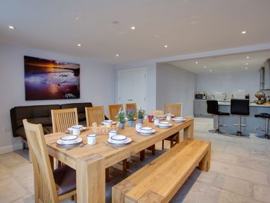 A dining area with a wooden table set with dishes and cups surrounded by wooden chairs and a bench beside a kitchen with stools at Low Tide in Weymouth