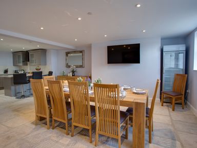 A dining area with a wooden table and chairs set with plates and cups next to a kitchen with two black bar stools at Low Tide in Weymouth
