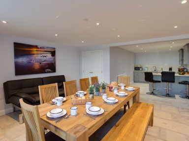 A dining area with a wooden table set with plates mugs and cutlery a black sofa in the background and a kitchen with bar stools at Low Tide in Weymouth