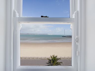 A window view showing a sandy beach a palm tree a road and the sea with a pier and distant hills at Low Tide in Weymouth
