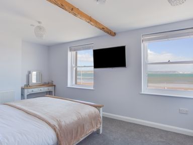 A bedroom with a bed a vanity table two windows showing a beach view and a wall-mounted television at Low Tide in Weymouth