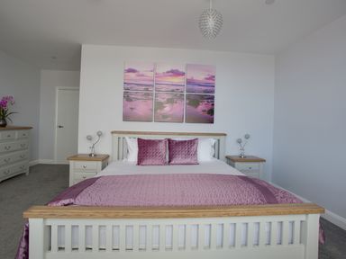A bedroom with a bed with purple pillows and blanket bedside tables and a dresser with flowers at Low Tide in Weymouth