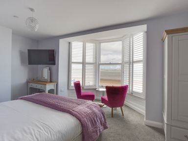 A bedroom with a bed a wall mounted TV two pink chairs by a window with shutters at Low Tide in Weymouth