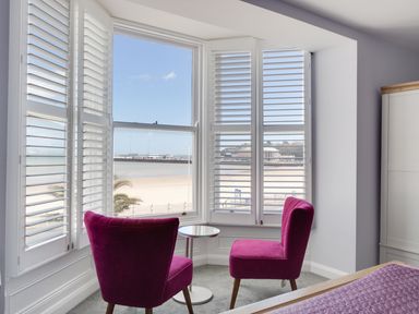 A corner of a room with two pink chairs and a small round table by windows with white shutters overlooking a beach at Low Tide in Weymouth