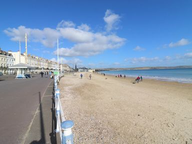 A beach with people walking along the shore and a row of buildings beside a promenade at Low Tide in Weymouth