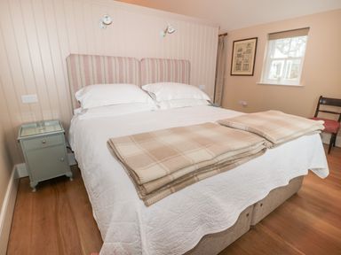 A bedroom with a double bed two beige blankets a bedside table and a chair near a window at The Garden Cottage in West Woodburn