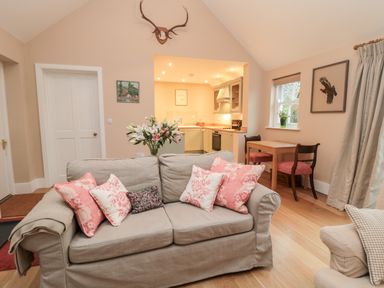 A living room with a sofa with pink cushions a small wooden table with two chairs and an open kitchen area at The Garden Cottage in West Woodburn