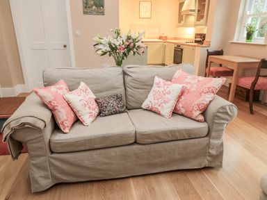A beige sofa with pink and patterned cushions in a room with a wooden floor and an open kitchen at The Garden Cottage in West Woodburn