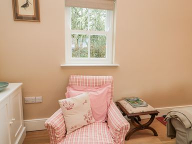 A plaid armchair with cushions by a window and a small wooden stool with a magazine at The Garden Cottage in West Woodburn