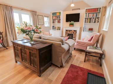 A living room with a sofa a wooden chest with flowers a fireplace with stacked firewood bookshelves and armchairs at The Garden Cottage in West Woodburn