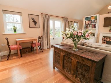 A living room with a wooden table and two chairs near a window and door a wooden chest with flowers on top a sofa a bookshelf and a fireplace at The Garden Cottage in West Woodburn