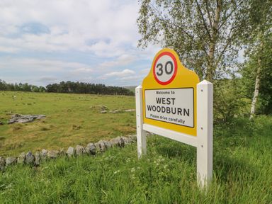 A yellow speed limit and welcome sign for West Woodburn beside grassy fields and trees at The Garden Cottage in West Woodburn
